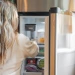 Back view of a blonde woman retrieving food from a refrigerator in a bright kitchen.